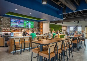 A bar area with multiple seating options, a television screen displaying sports, and a variety of bottles on shelves. - Candlewood Suites Kearney by IHG (Kearney)