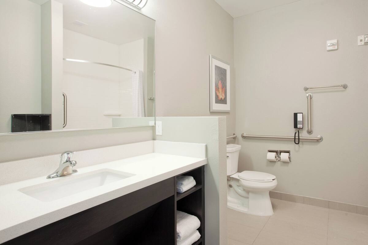 Un baño moderno con lavamanos, inodoro y ducha blancos. Se observan barras de apoyo y un teléfono fijados a la pared.