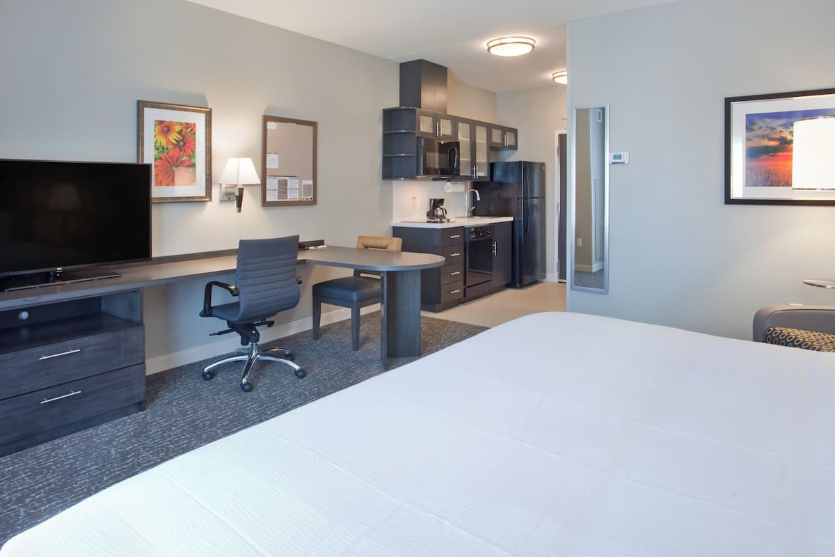 Habitación de hotel con una cama grande, un escritorio con silla, televisor, kitchenette y obras de arte en las paredes.