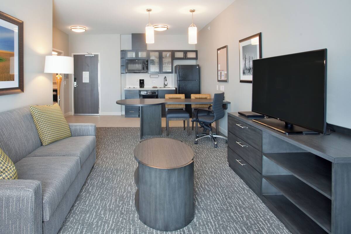 Una habitación de hotel moderna con un sofá gris, una mesa redonda, un televisor de pantalla plana, un escritorio con una silla, una zona de cocina y un ventanal con vistas a un campo.