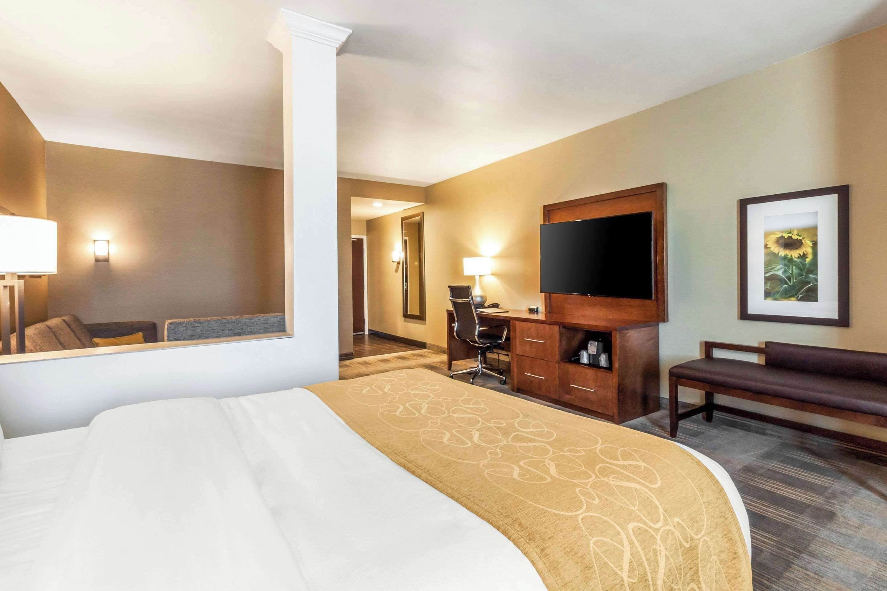 A hotel room with a large bed, a desk with a chair, a television, a bench, and a framed picture on the wall.