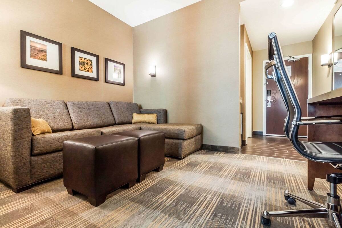 A hotel room with a sectional sofa, a small ottoman, a desk with a chair, framed pictures on the wall, and a carpeted floor.