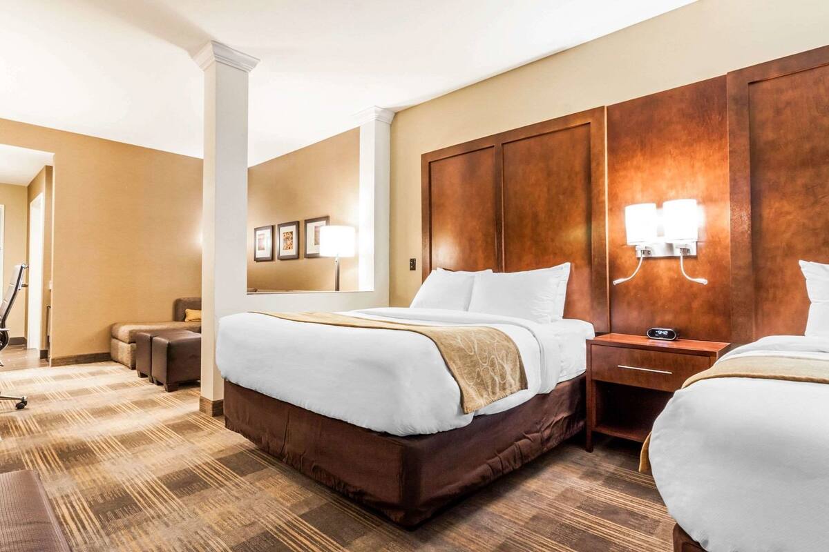 A hotel room with a large bed, a smaller bed, a desk, a chair, a lamp, and a wall-mounted TV.