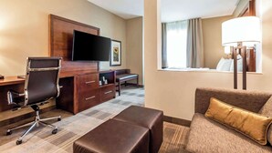 Une chambre d’hôtel équipée d’une télévision à écran plat, d’un bureau en bois, d’un fauteuil en cuir, d’un canapé agrémenté d’un coussin doré et d’une grande fenêtre avec des rideaux.