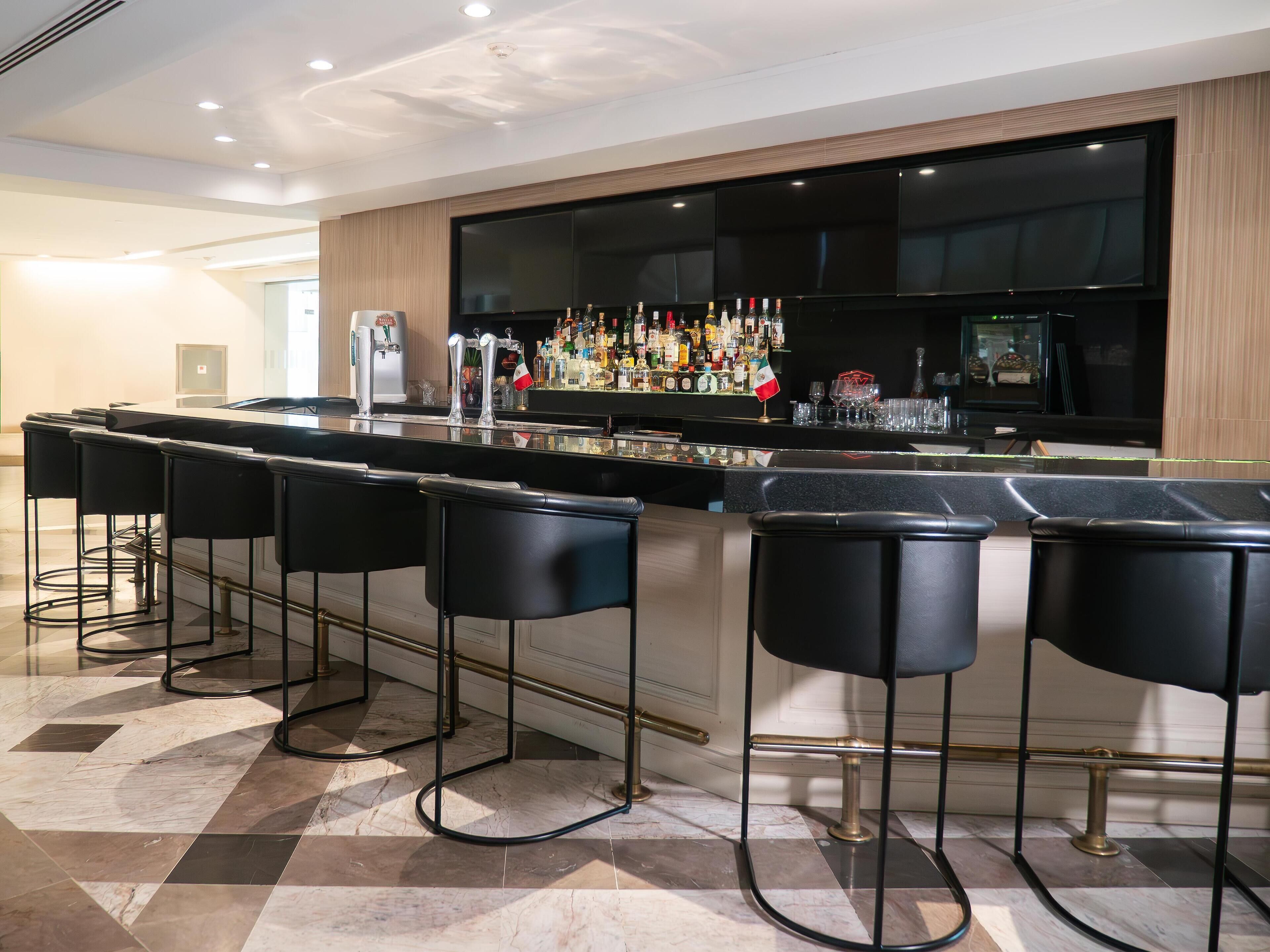 Zona de bar moderna con una barra alargada, varios taburetes y una amplia variedad de botellas y vasos.