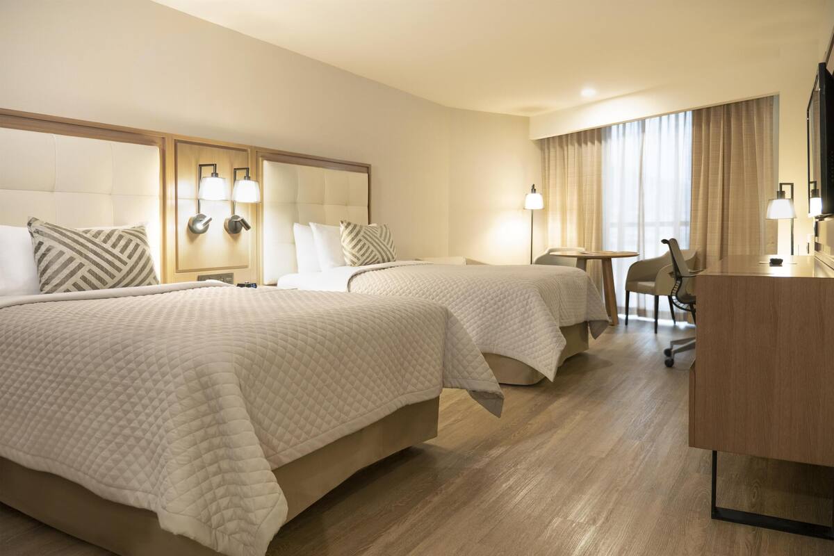 Ett hotellrum med två sängar, ett skrivbord, en stol och ett stort fönster med gardiner.