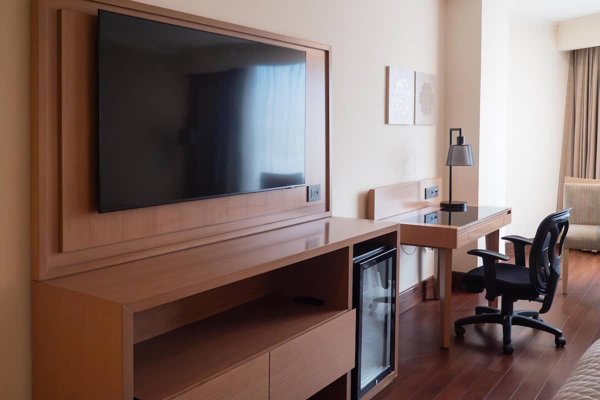 Ett hotellrum med en stor platt-TV som är monterad på väggen, ett skrivbord med en stol, ett litet kylskåp och en soffa.