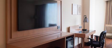 Habitación de hotel con una televisión de pantalla plana grande montada en la pared, un escritorio con una silla, un refrigerador pequeño y un sofá.