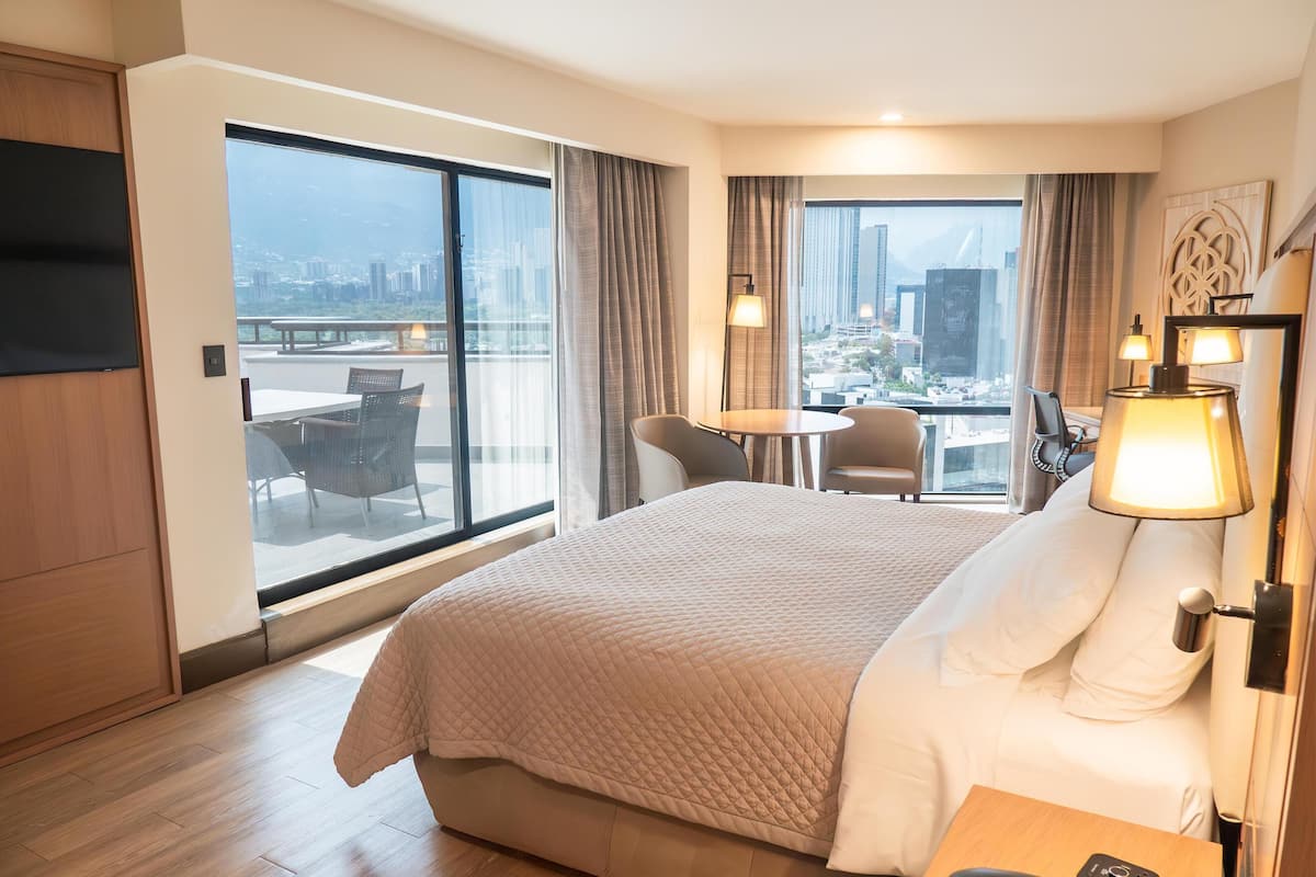 Ett hotellrum med en stor säng, ett skrivbord med en telefon, en stol, ett bord och utsikt över staden genom stora fönster.