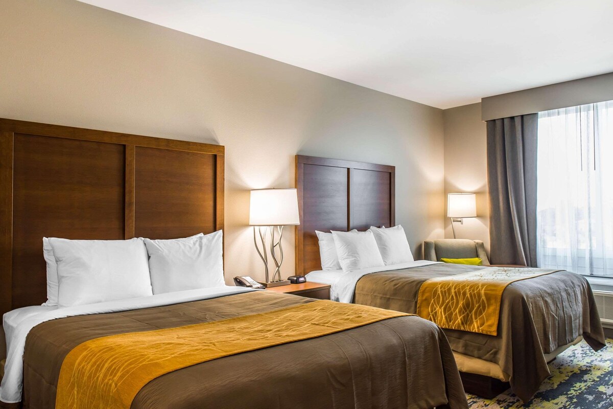 A hotel room with two beds, each with a wooden headboard, white bedding, and a brown throw blanket. There are bedside lamps and a window with curtains.