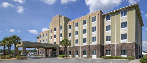A multi-story hotel with a covered entrance, parking lot, and a clear sky.