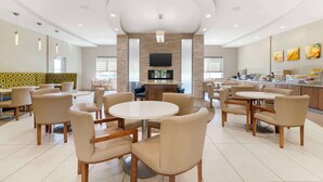 A spacious dining area with round tables and chairs, a flat-screen TV mounted on the wall, and a kitchen area visible in the background.
