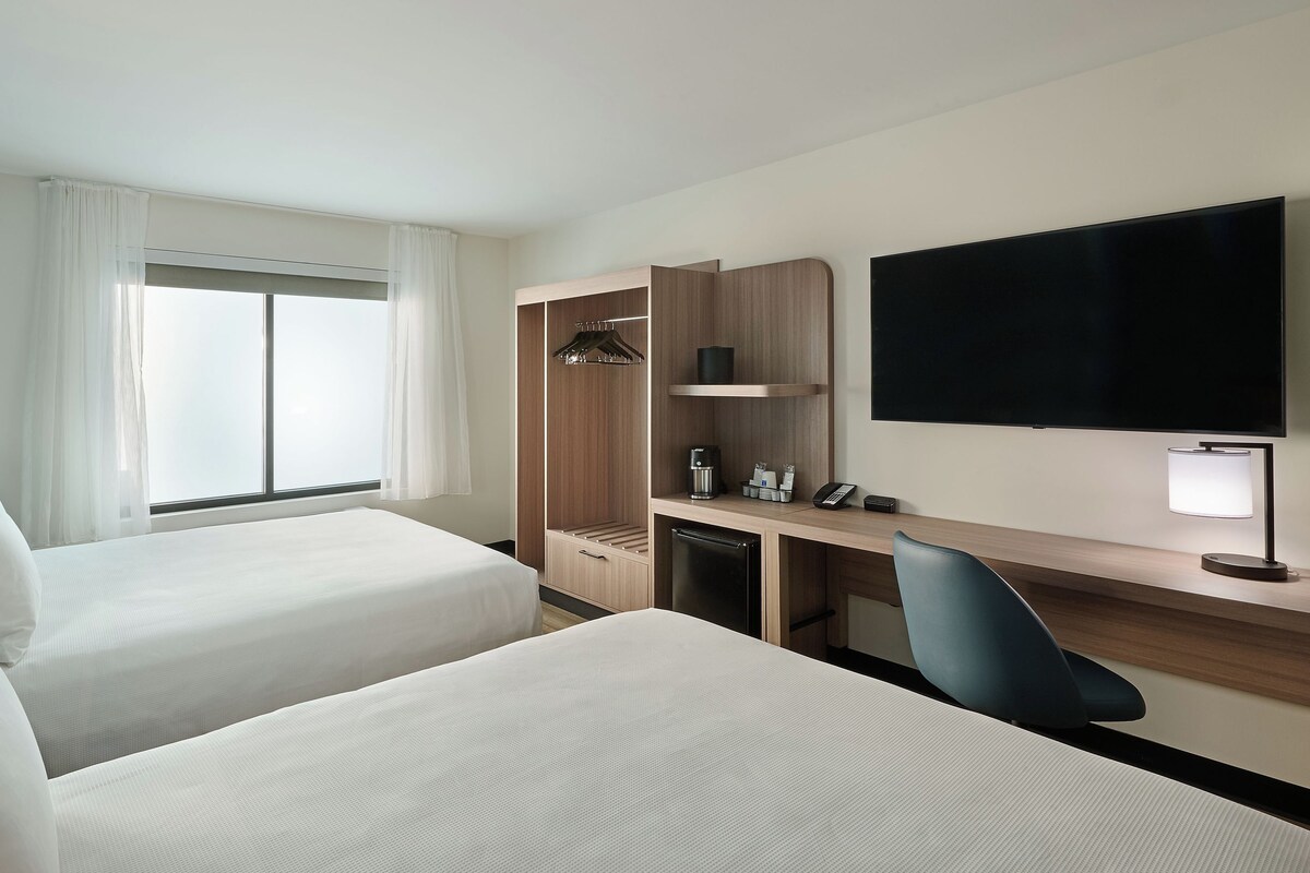 A hotel room with two beds, a desk, a TV, a wardrobe, and a window with curtains.