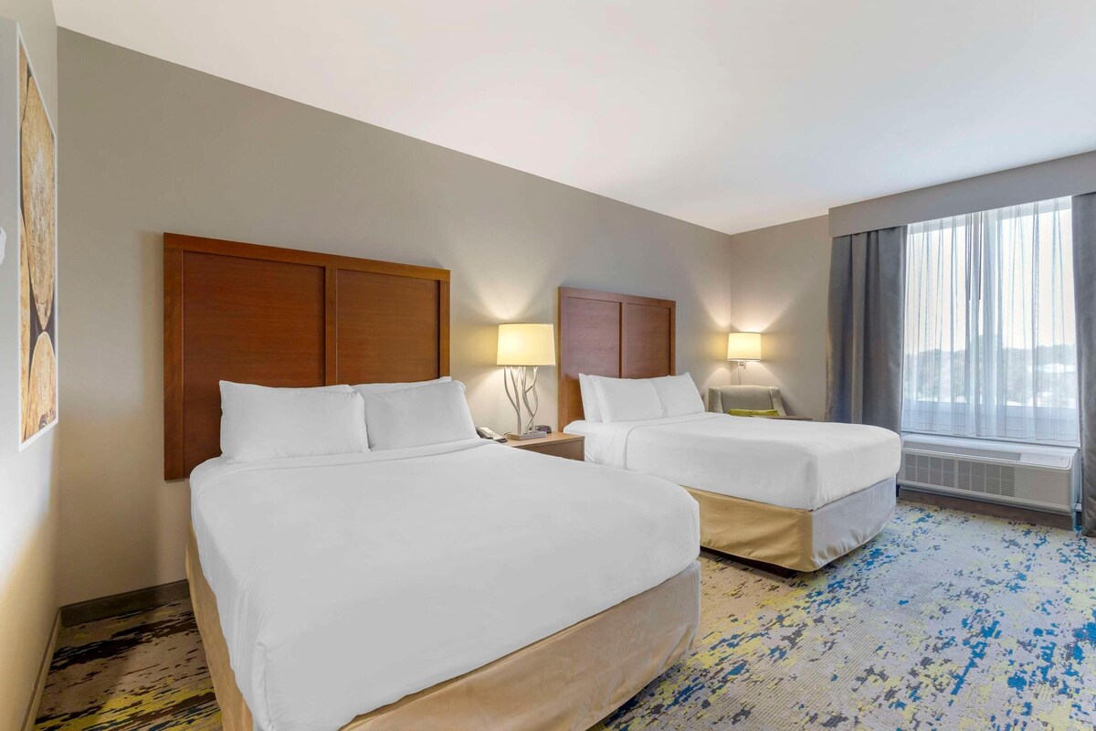 A hotel room with two beds, wooden headboards, bedside lamps, a large window with curtains, and a patterned carpet.