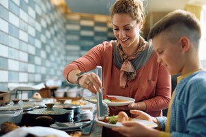 Una persona sirve comida a otra en una cocina con paredes de azulejos y diversos utensilios de cocina.