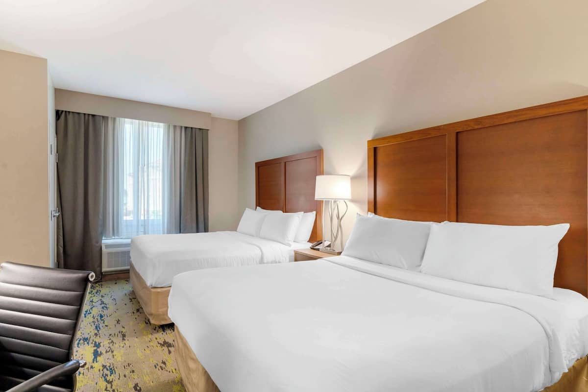 Ein Hotelzimmer mit zwei Betten, einem Holzkopfteil, einem Schreibtisch mit Lampe, einem Stuhl und einem Vorhangfenster.
