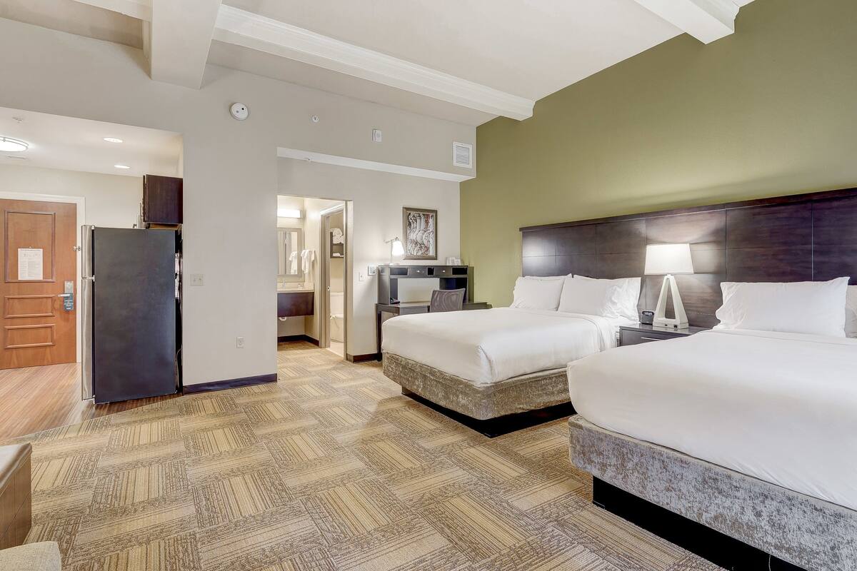 A hotel room with two beds, a desk, a refrigerator, and a bathroom visible through an open door.