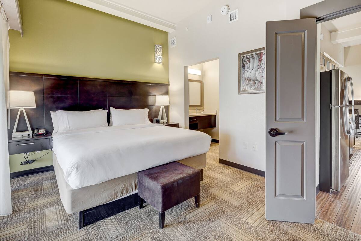 A hotel room with a large bed, bedside tables, a bench, a door, and a view into the bathroom.