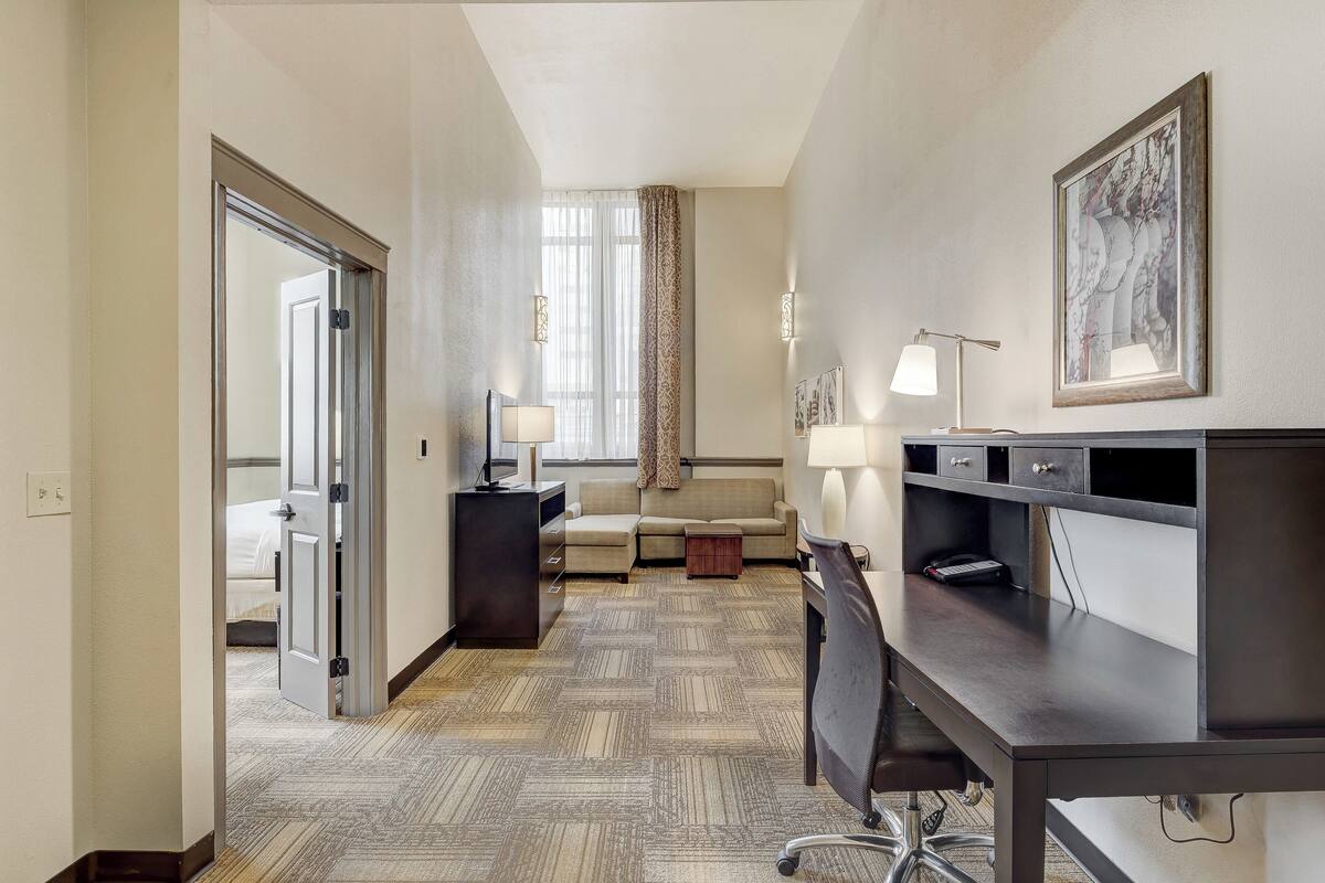 A hotel room with a bed, a desk, a chair, a sofa, a small table, a lamp, and a framed picture on the wall.