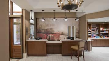A hotel lobby with a reception desk, a chandelier, pendant lights, a large abstract painting, and a retail area with shelves stocked with products.