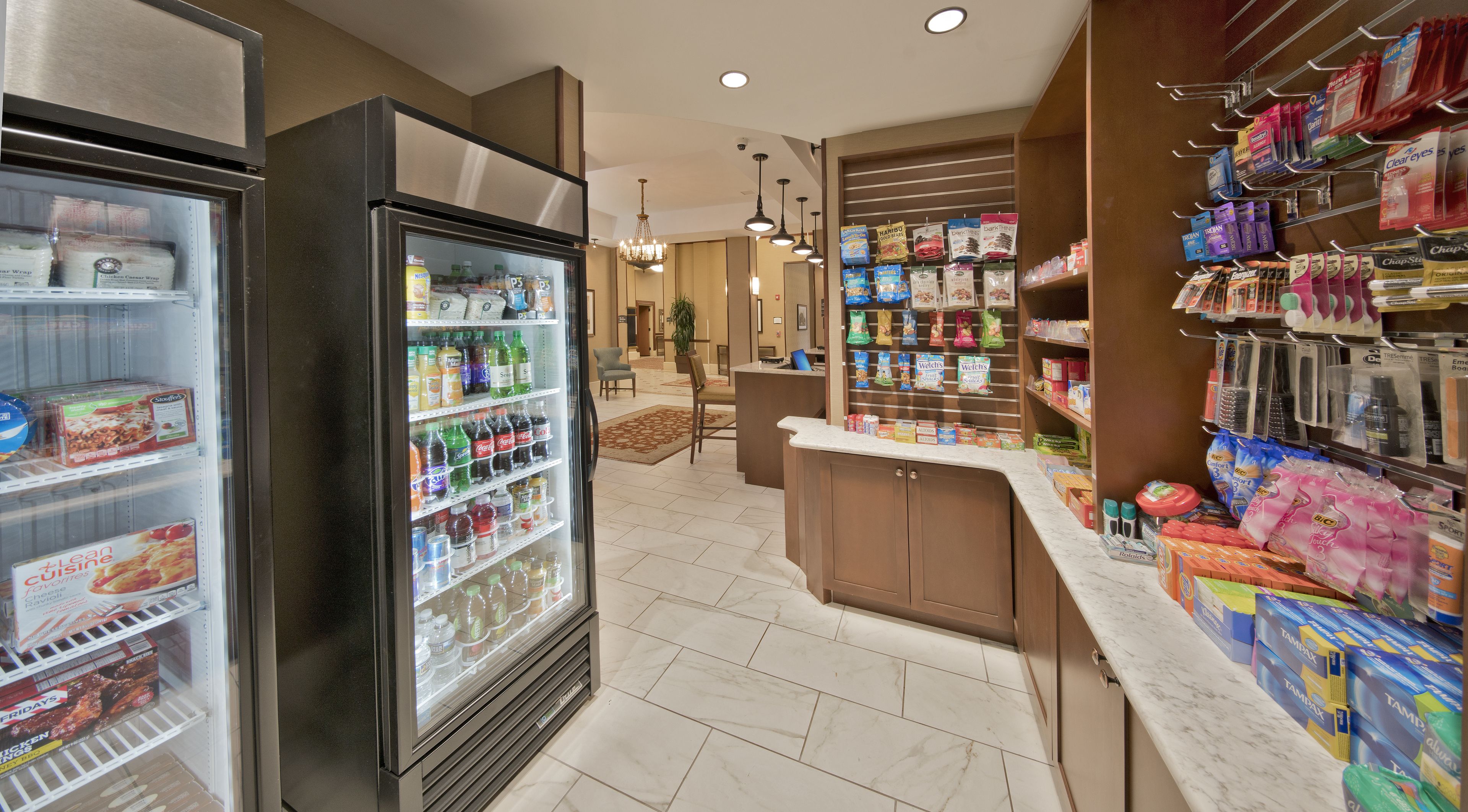 A hotel lobby with a self-service beverage fridge, a reception desk, and a variety of snacks and toiletries on shelves.