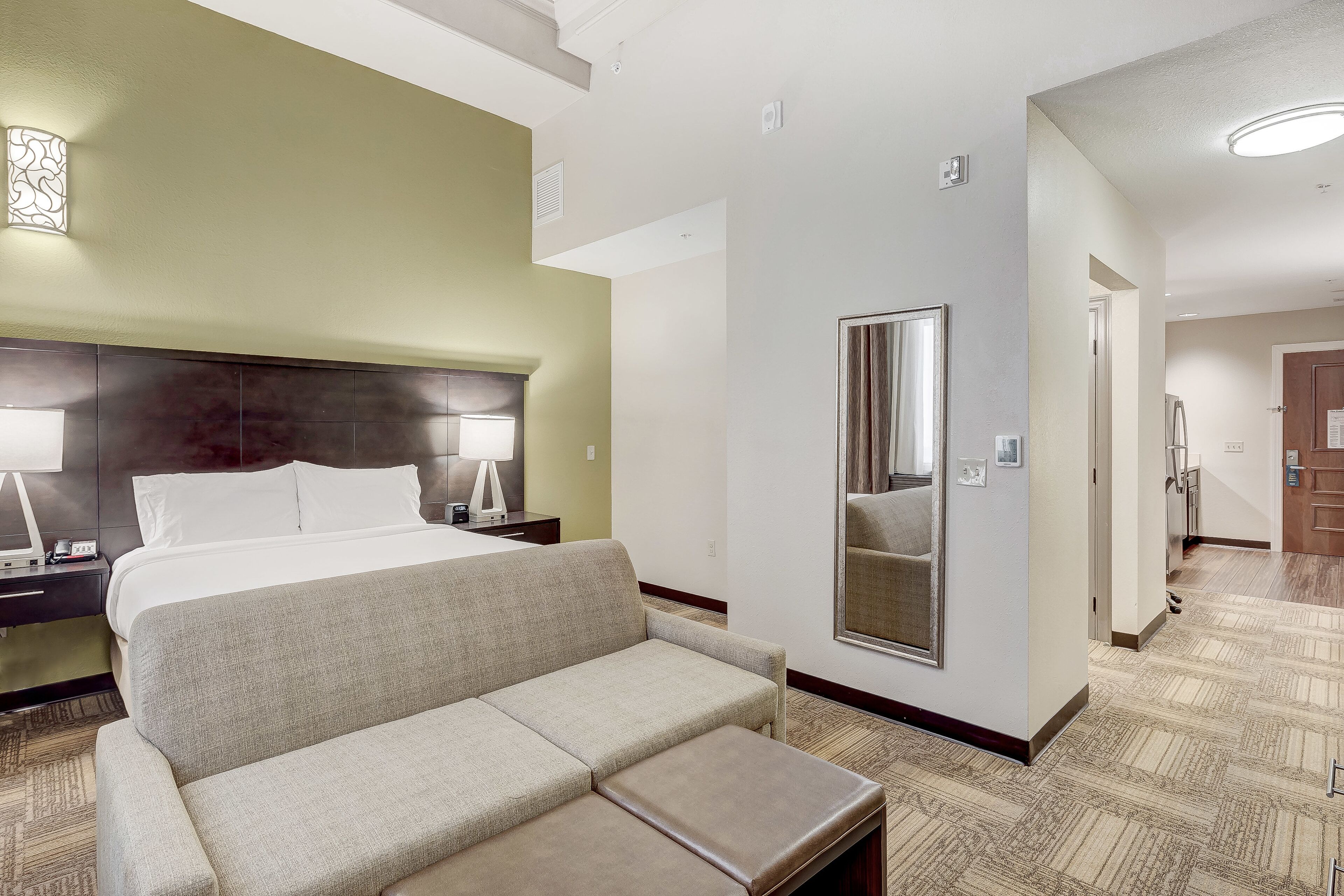 A hotel room with a bed, a sofa, a nightstand, a lamp, a mirror, and a carpeted floor.