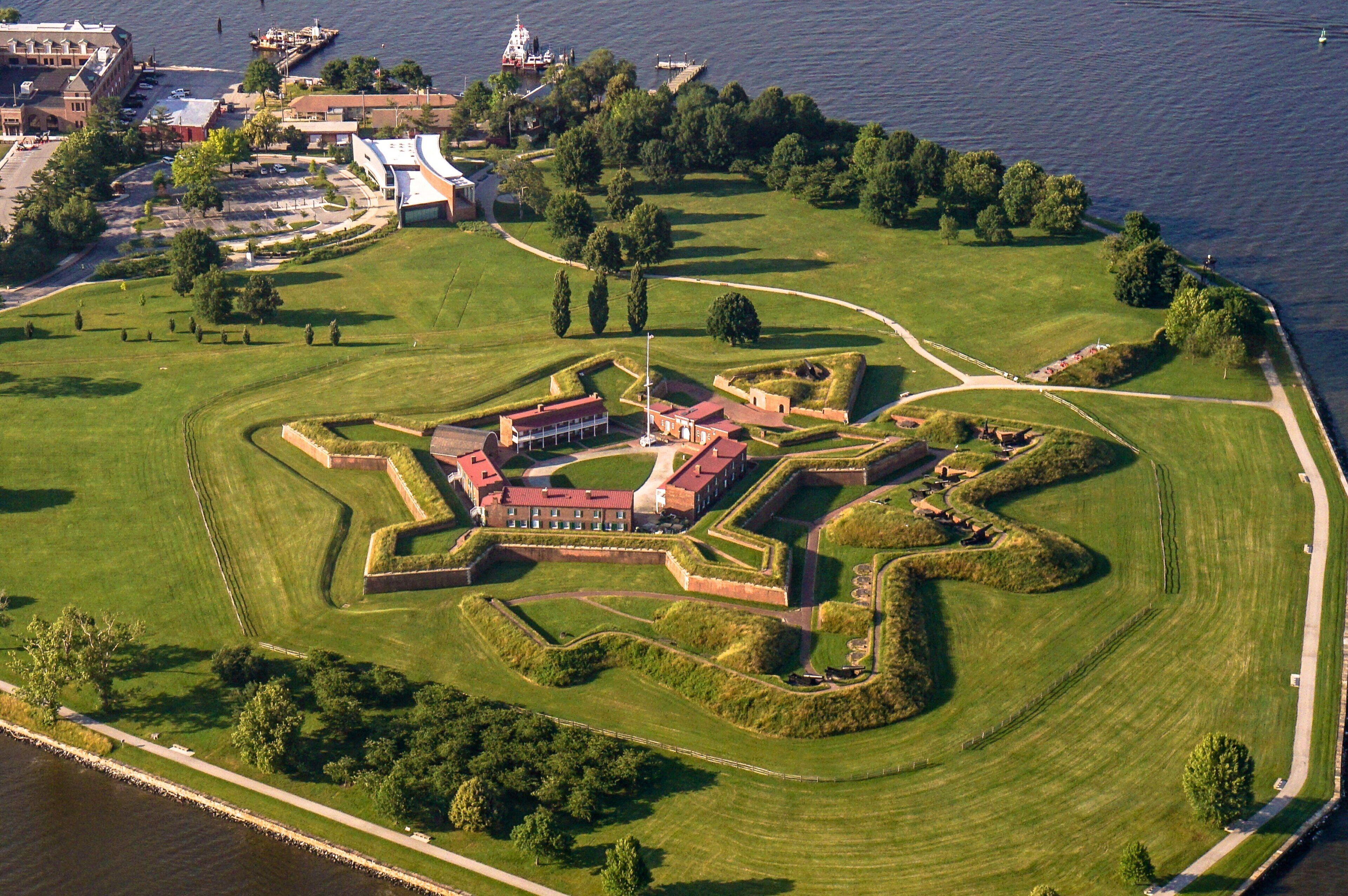 Eine Vogelperspektive zeigt eine historische Festung mit einem zentralen Gebäude, umgeben von gepflegten Rasenflächen und einem Gewässer.