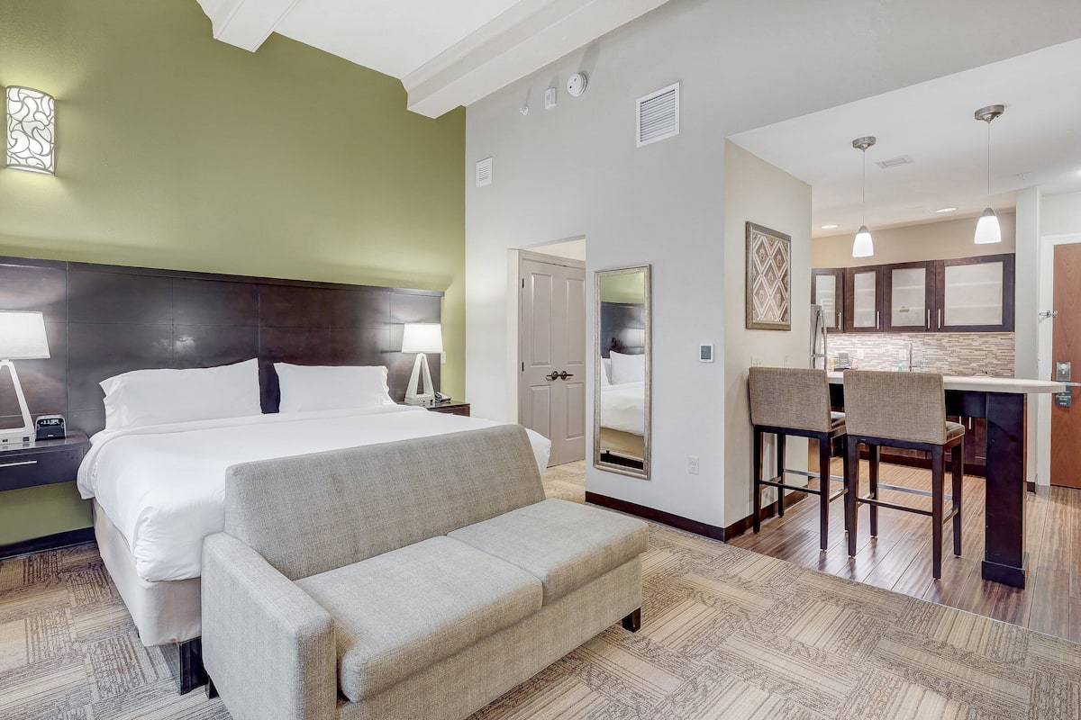 A hotel room with a bed, a sofa, a desk with two chairs, a mirror, and a kitchenette.