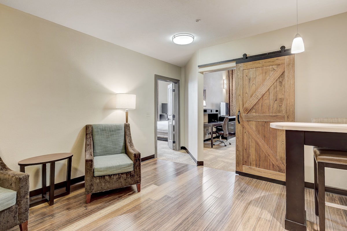 A hotel room with a wooden floor, a sliding barn door, a small table with a lamp, and a chair.