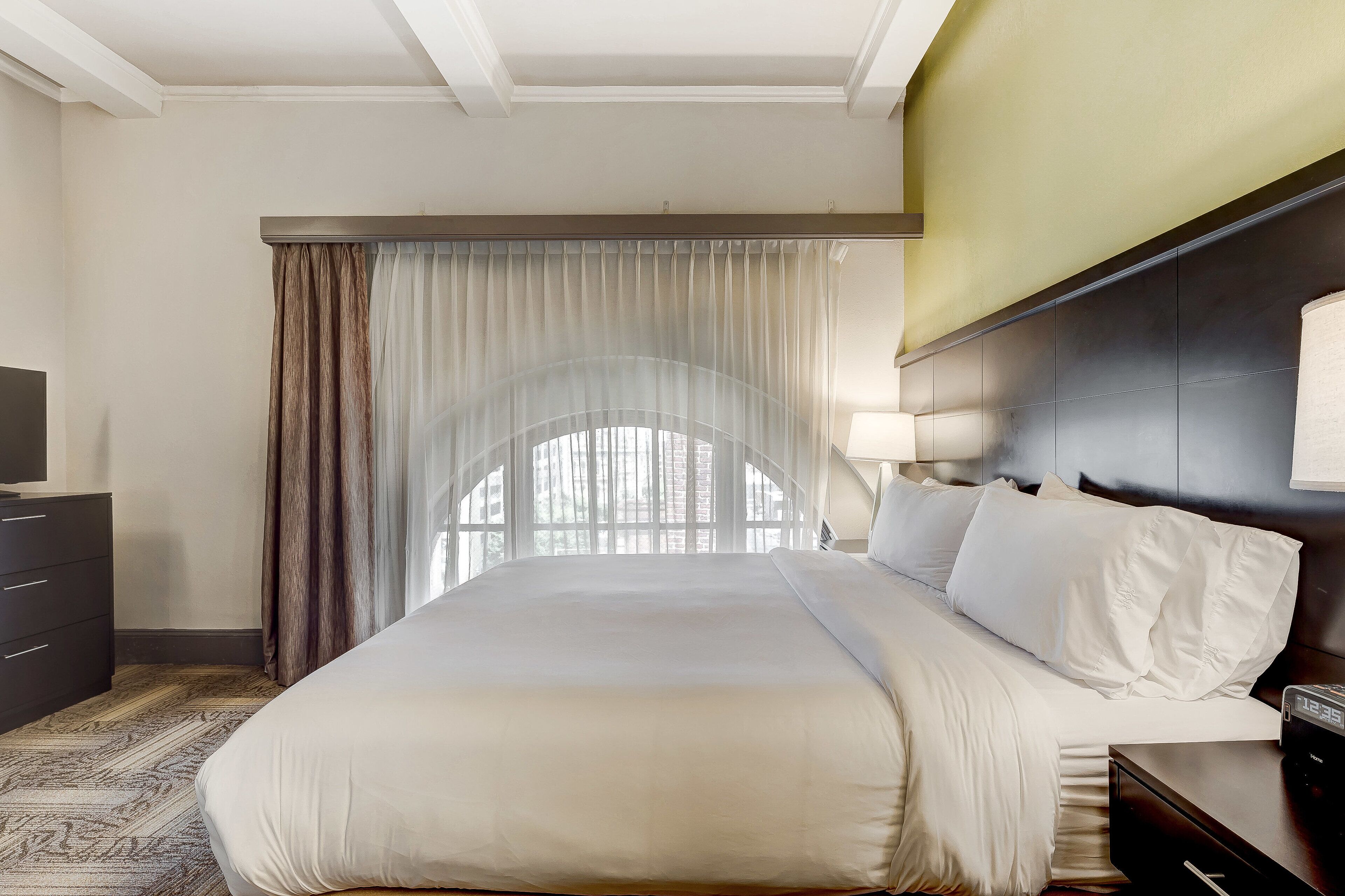Habitación de hotel con una cama grande, televisión, escritorio y vista a árboles a través de cortinas translúcidas.