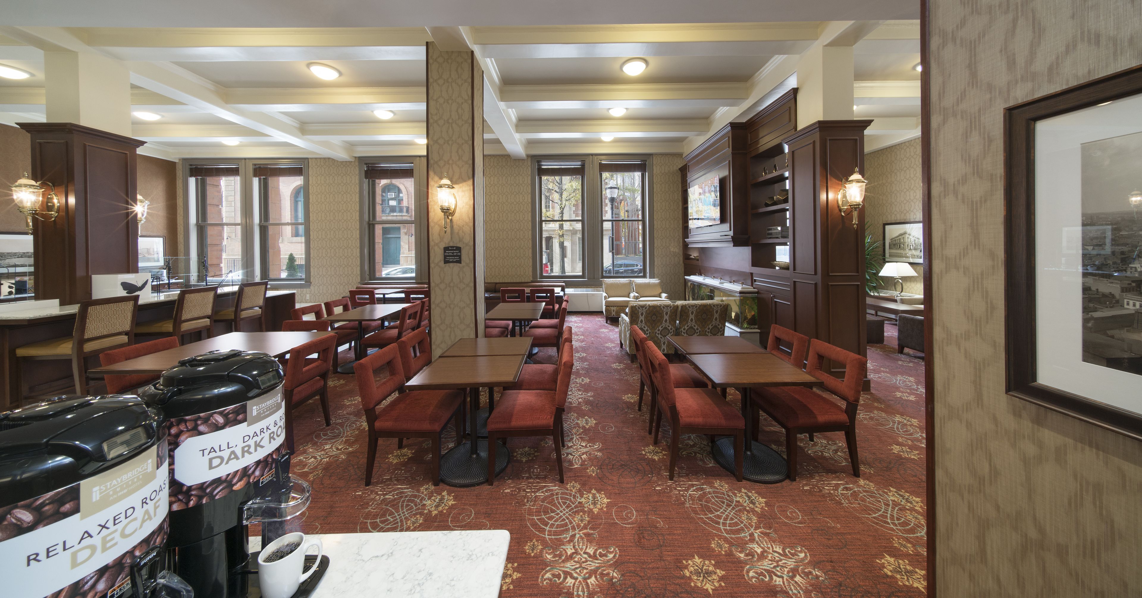 A hotel dining area with multiple tables and chairs, a coffee station with decaf and dark roast options, and a view of the street through large windows.