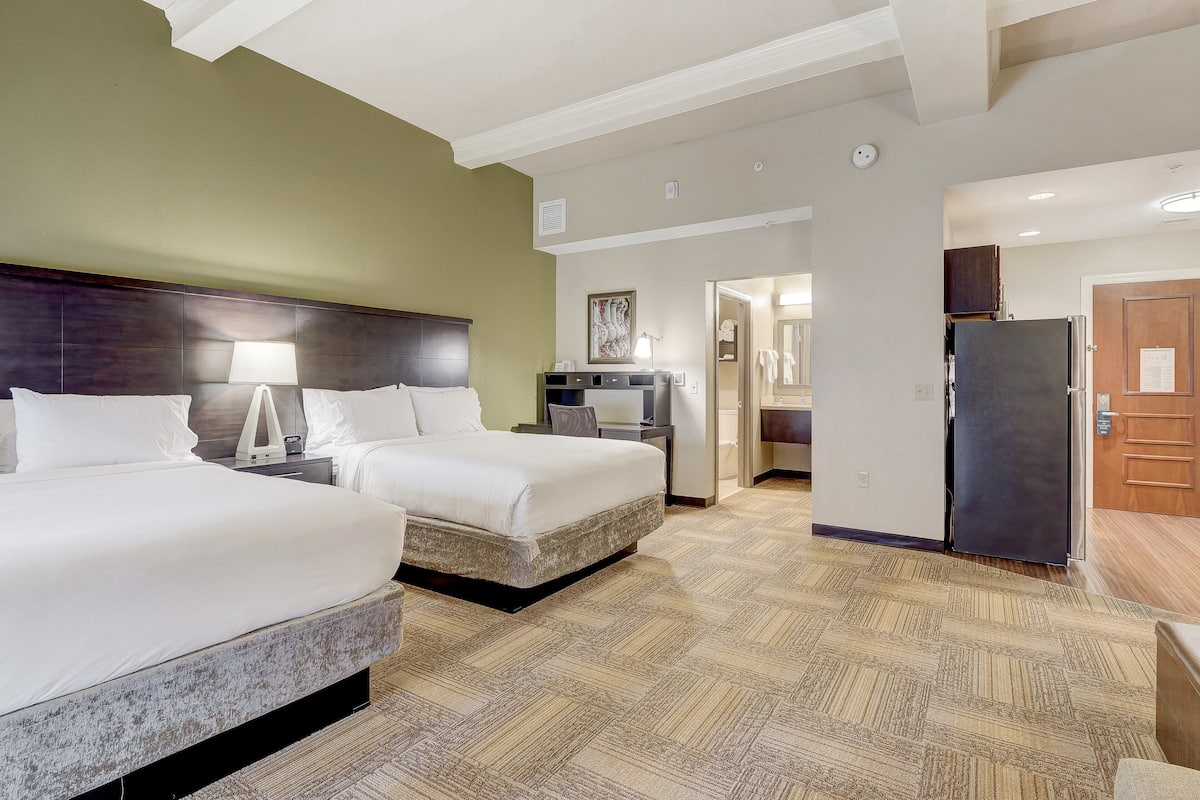 A hotel room with two beds, a desk, a chair, a lamp, a refrigerator, and a door leading to a bathroom.