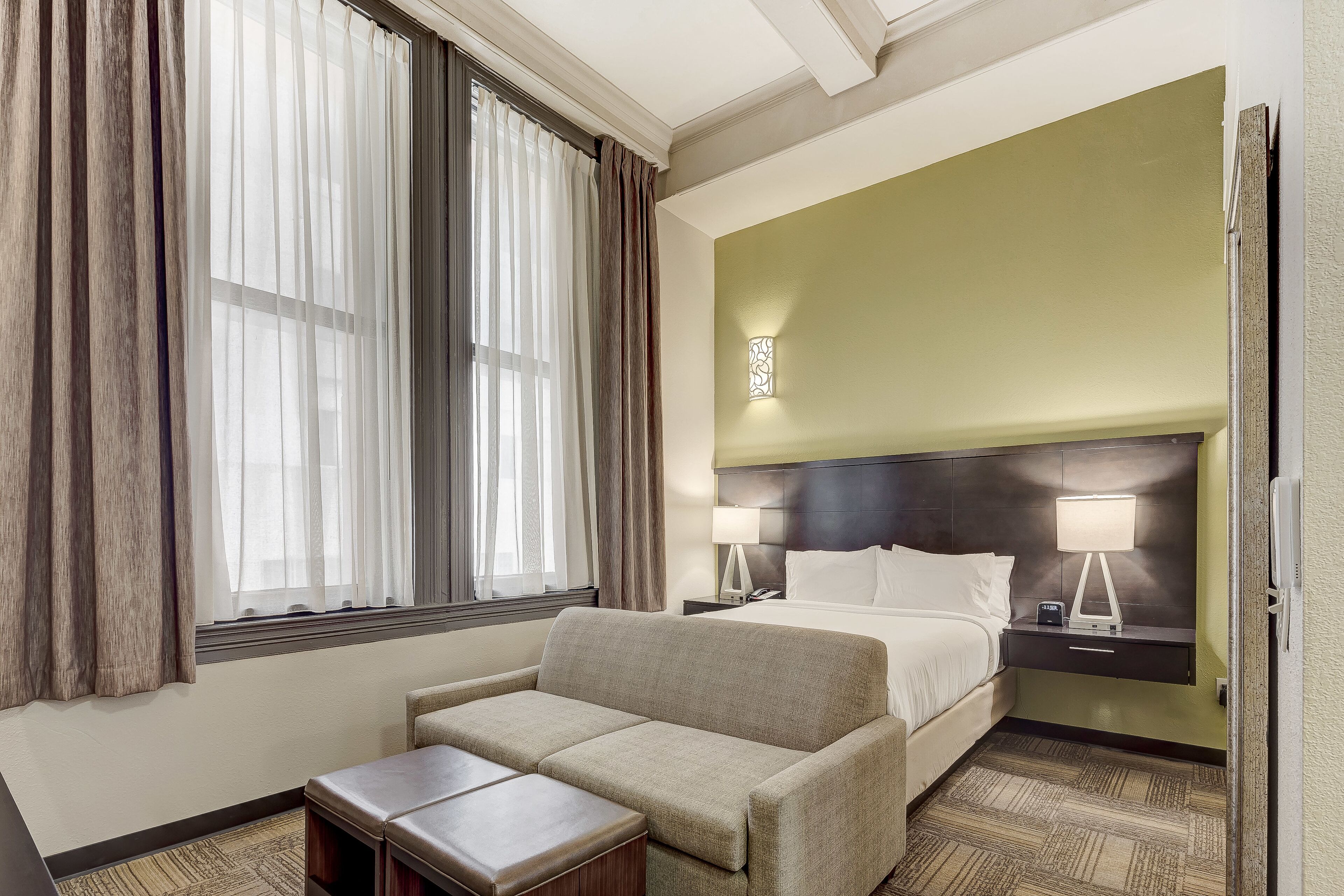 A hotel room with a bed, a sofa, a nightstand with a lamp, and a window with curtains.