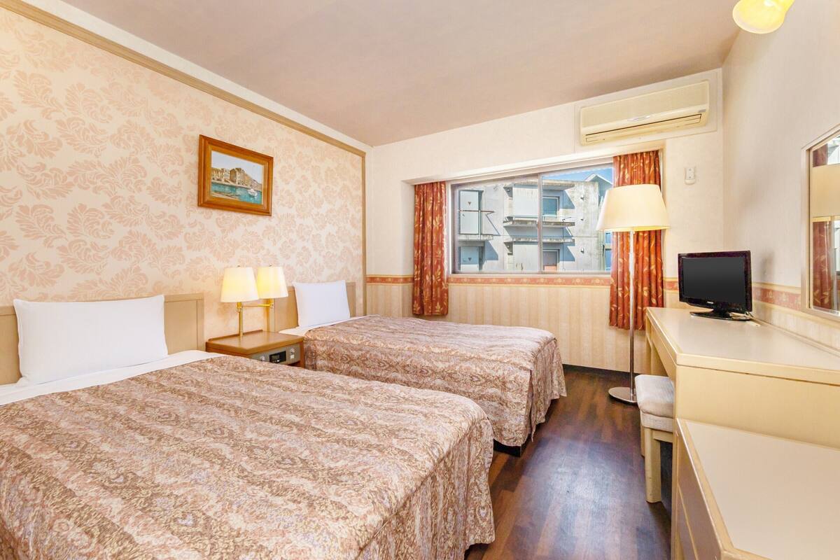 Habitación de hotel con dos camas, un escritorio con computadora, aire acondicionado y ventana con vista a edificios.