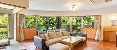 A spacious living room with a large sectional sofa, a wooden coffee table, and a balcony with a view of greenery.