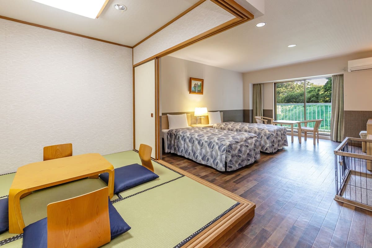 Habitación de hotel con zona de estar con esterilla de tatami, dos camas con ropa de cama estampada, un escritorio con una silla, un balcón con vistas a zonas verdes y un cuadro en la pared.
