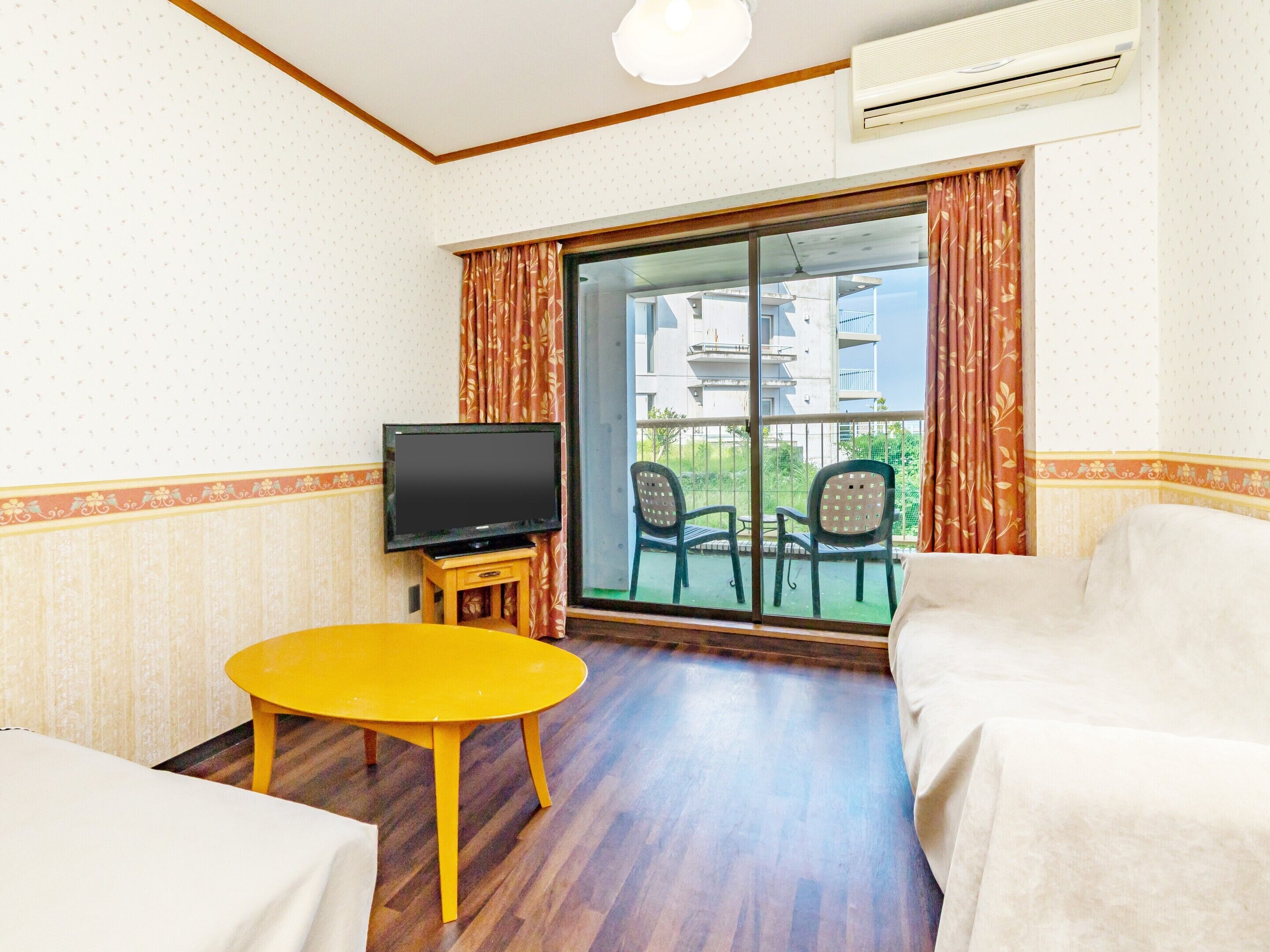 A hotel room with a television on a wooden stand, a yellow table, two white armchairs, and a large sliding glass door leading to an outdoor balcony with chairs.