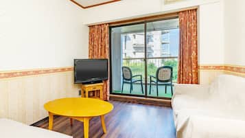 A hotel room with a television on a wooden stand, a yellow table, two white armchairs, and a large sliding glass door leading to an outdoor balcony with chairs.