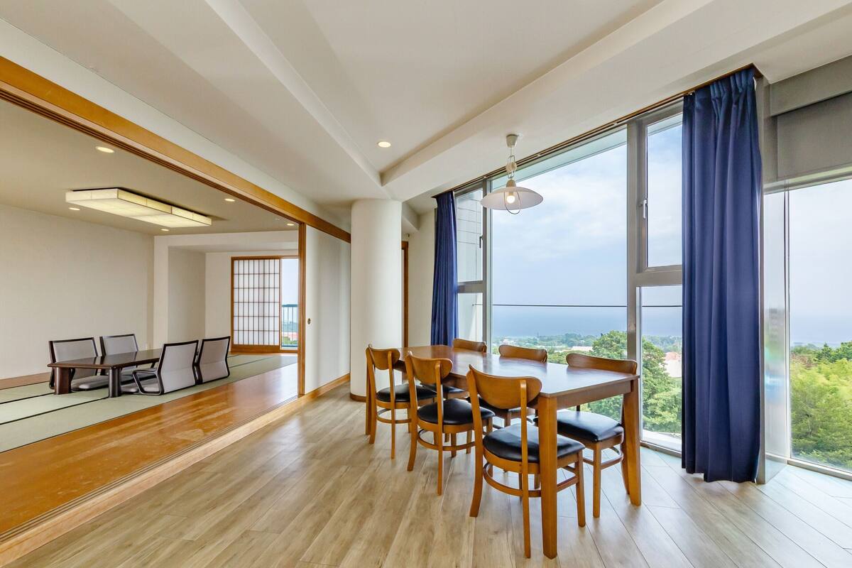 Un amplio comedor con una mesa y sillas de madera, ventanas grandes con cortinas azules y vista a la vegetación.