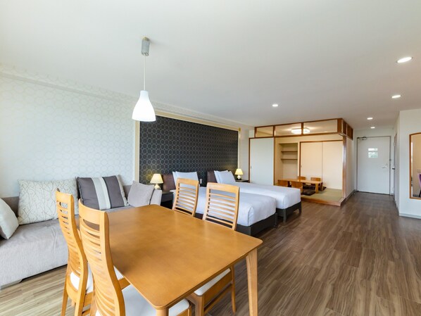 A modern hotel room with a wooden dining table, two beds, a sofa, and a separate dining area.