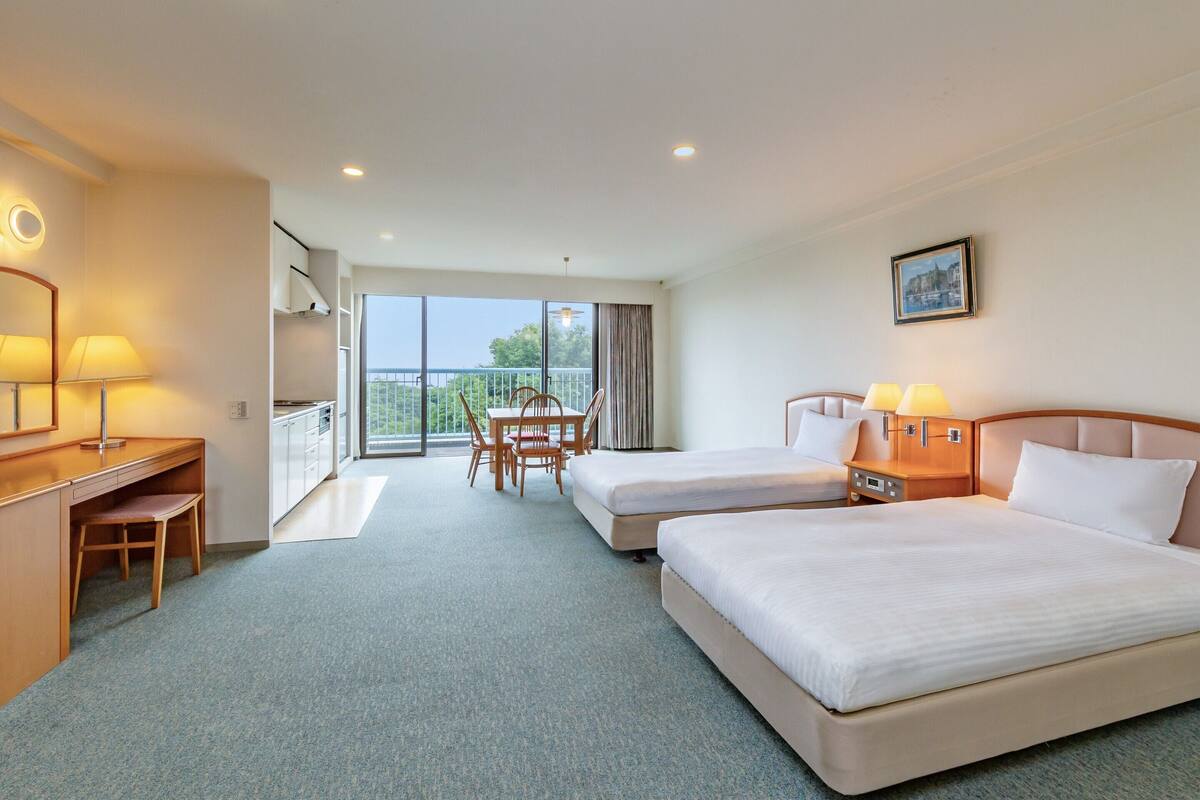 Habitación de hotel con dos camas, un escritorio, una silla, un balcón con vistas y un cuadro en la pared.