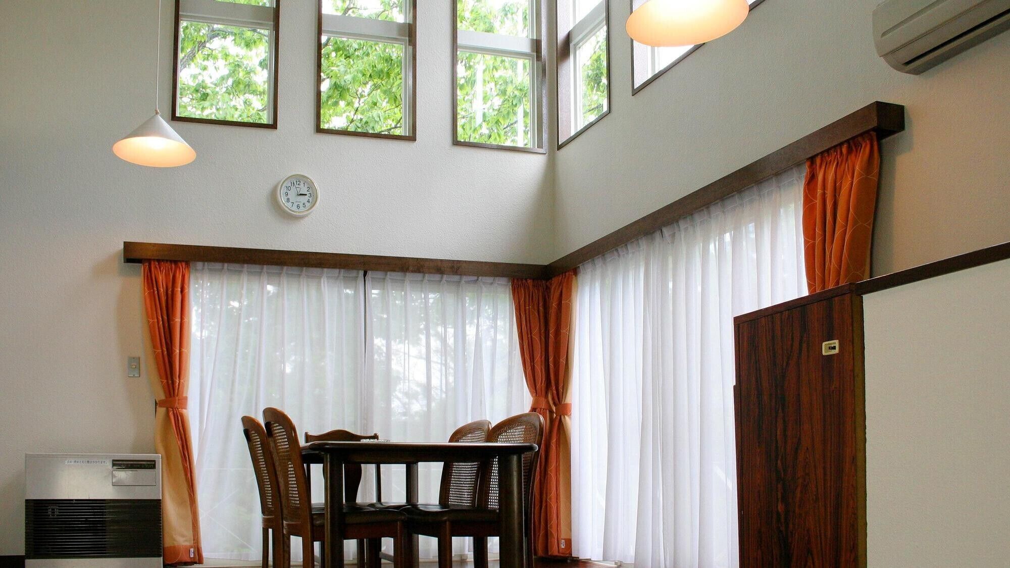 A dining area with a table and chairs, large windows with curtains, an air conditioning unit, and a clock on the wall.