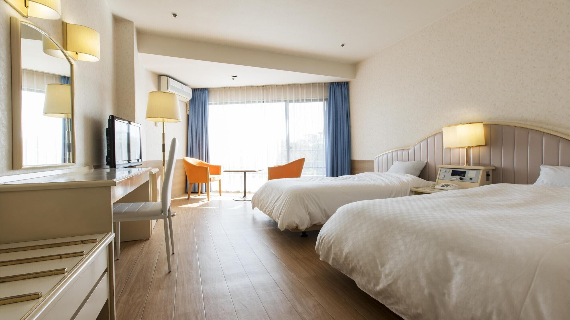 Ein Hotelzimmer mit zwei Betten, einem Schreibtisch mit Fernseher, einem Sessel, einem kleinen Tisch und einem Fenster mit Vorhängen.