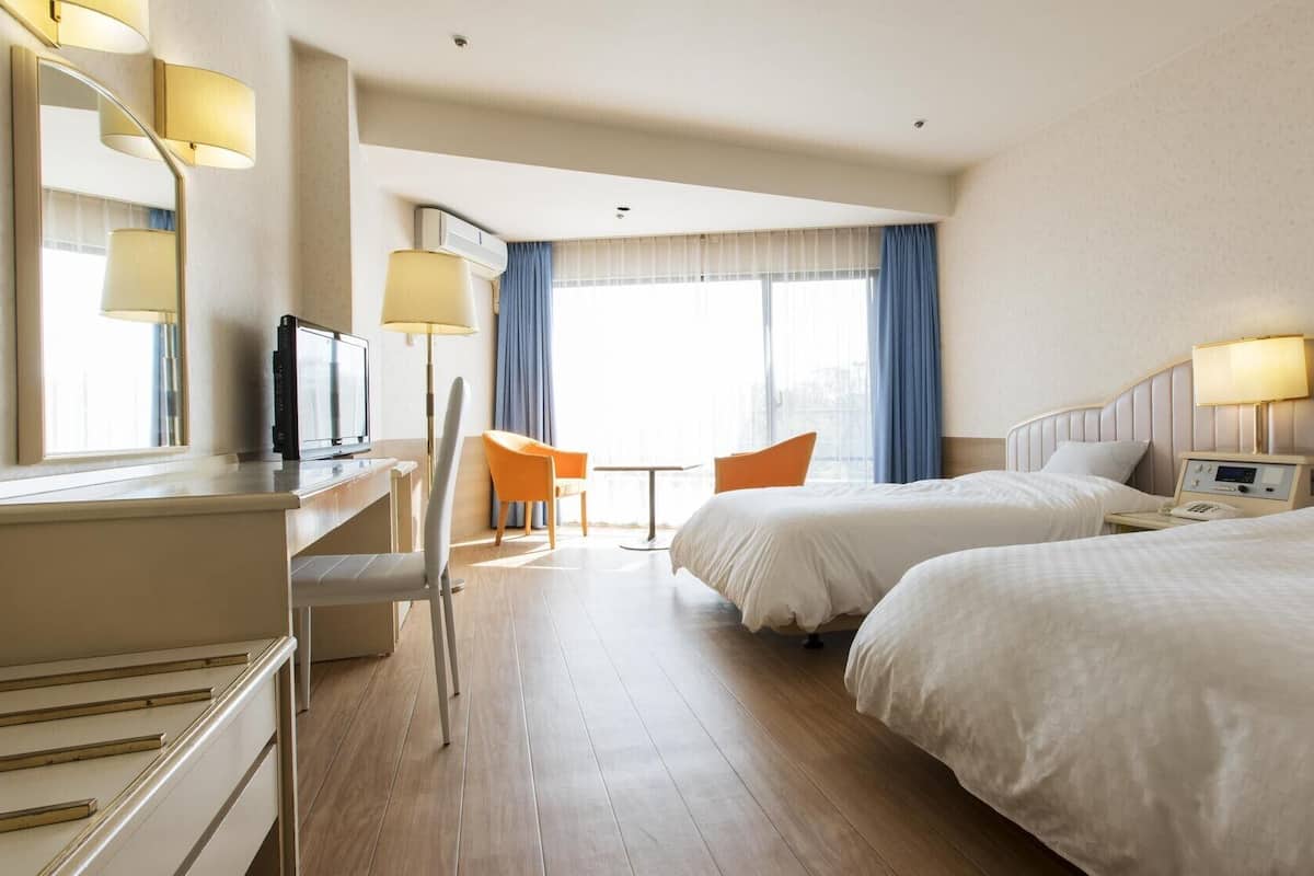 A hotel room with two beds, a desk with a TV, a chair, a small table, and a window with curtains.