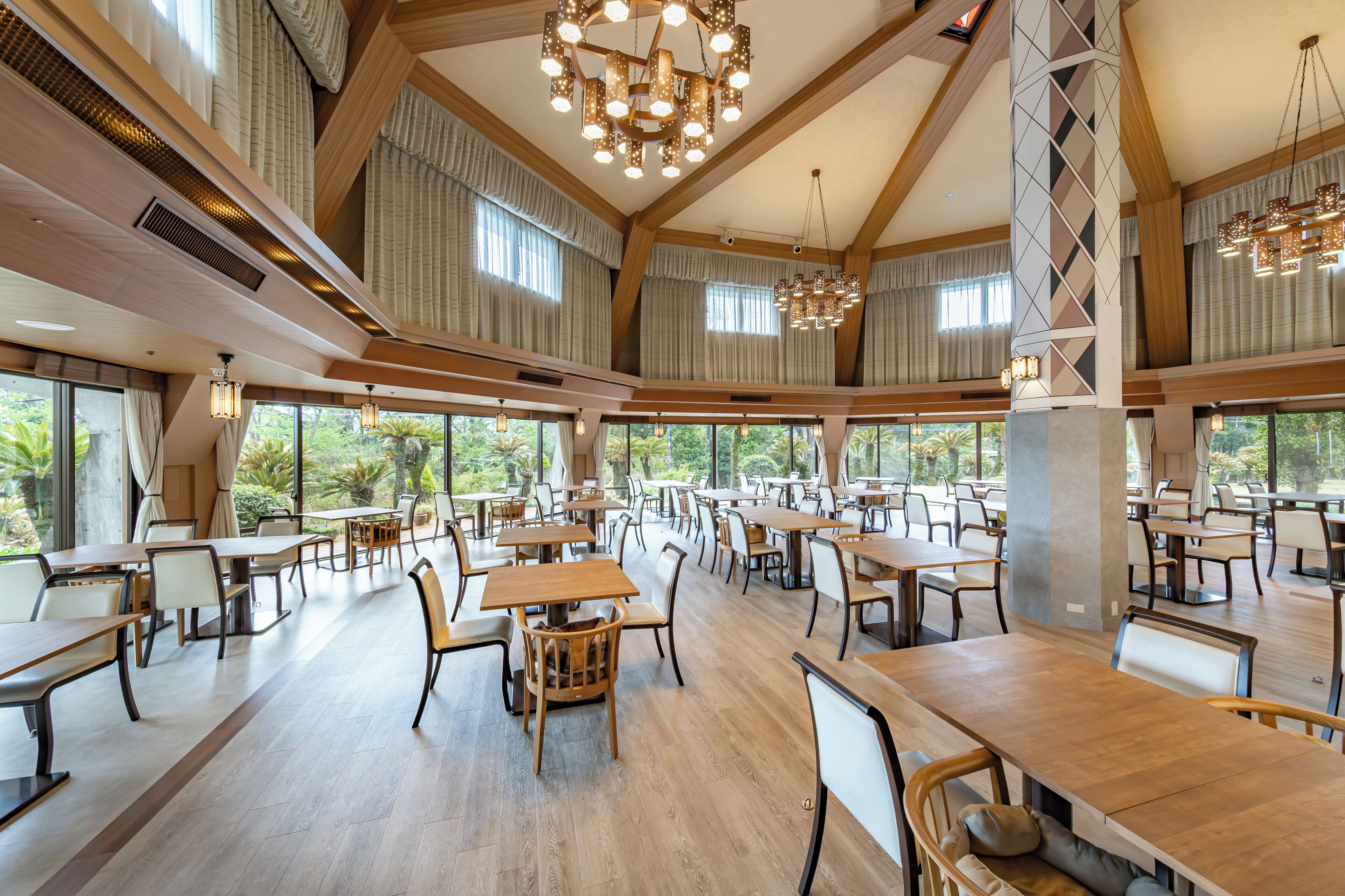 A spacious dining area with multiple tables and chairs, large windows offering views of the outdoors, and a high ceiling with wooden beams and modern lighting fixtures.