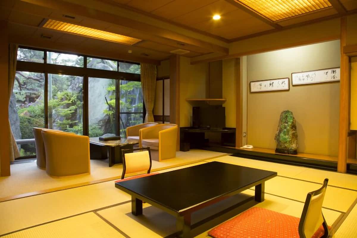 Une pièce avec un sol recouvert de tatamis, comprenant une table basse en tatami, des coussins, une cheminée et une vue sur la verdure environnante.