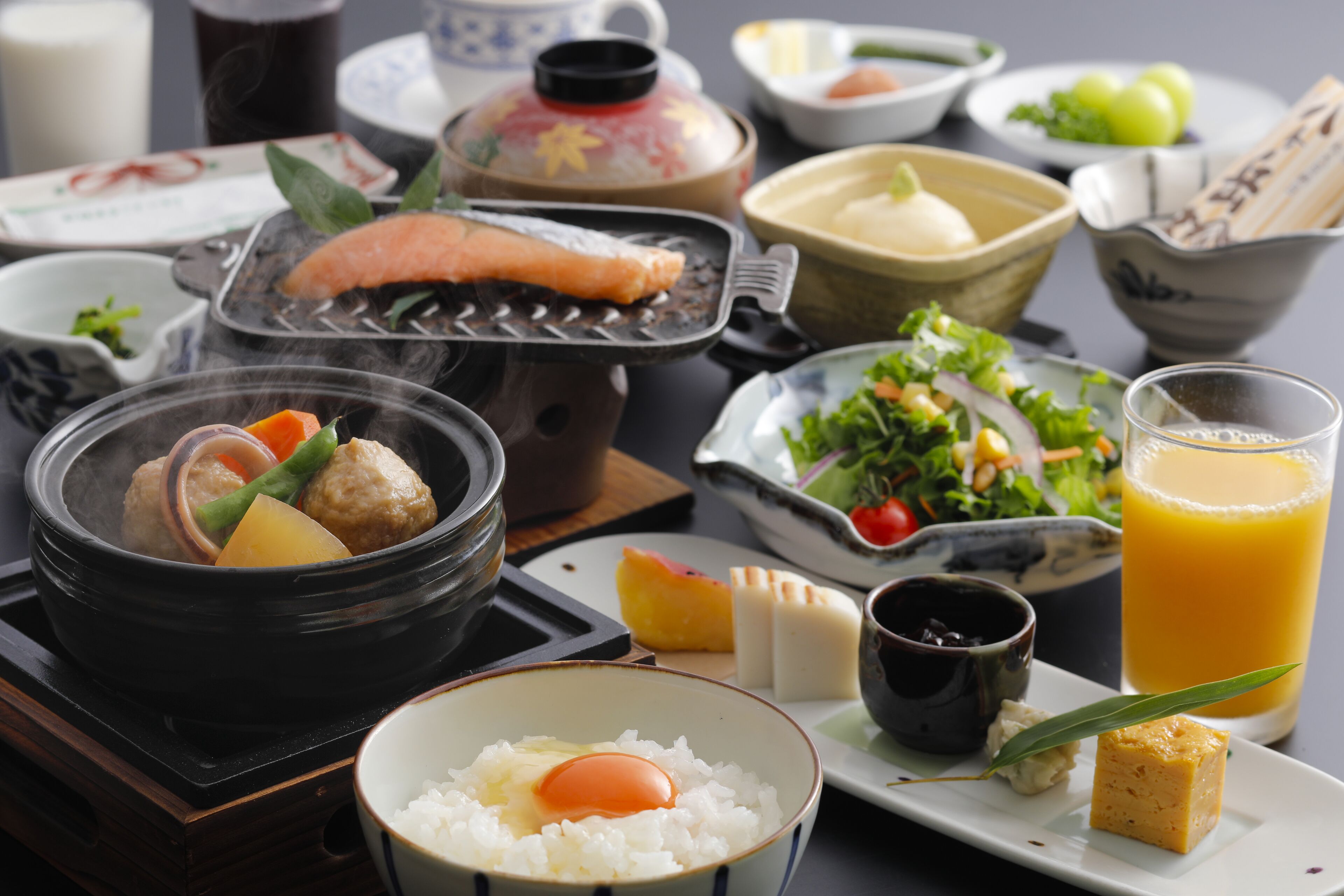 A variety of dishes including a hot pot, rice bowl with an egg, a glass of orange juice, and a salad.