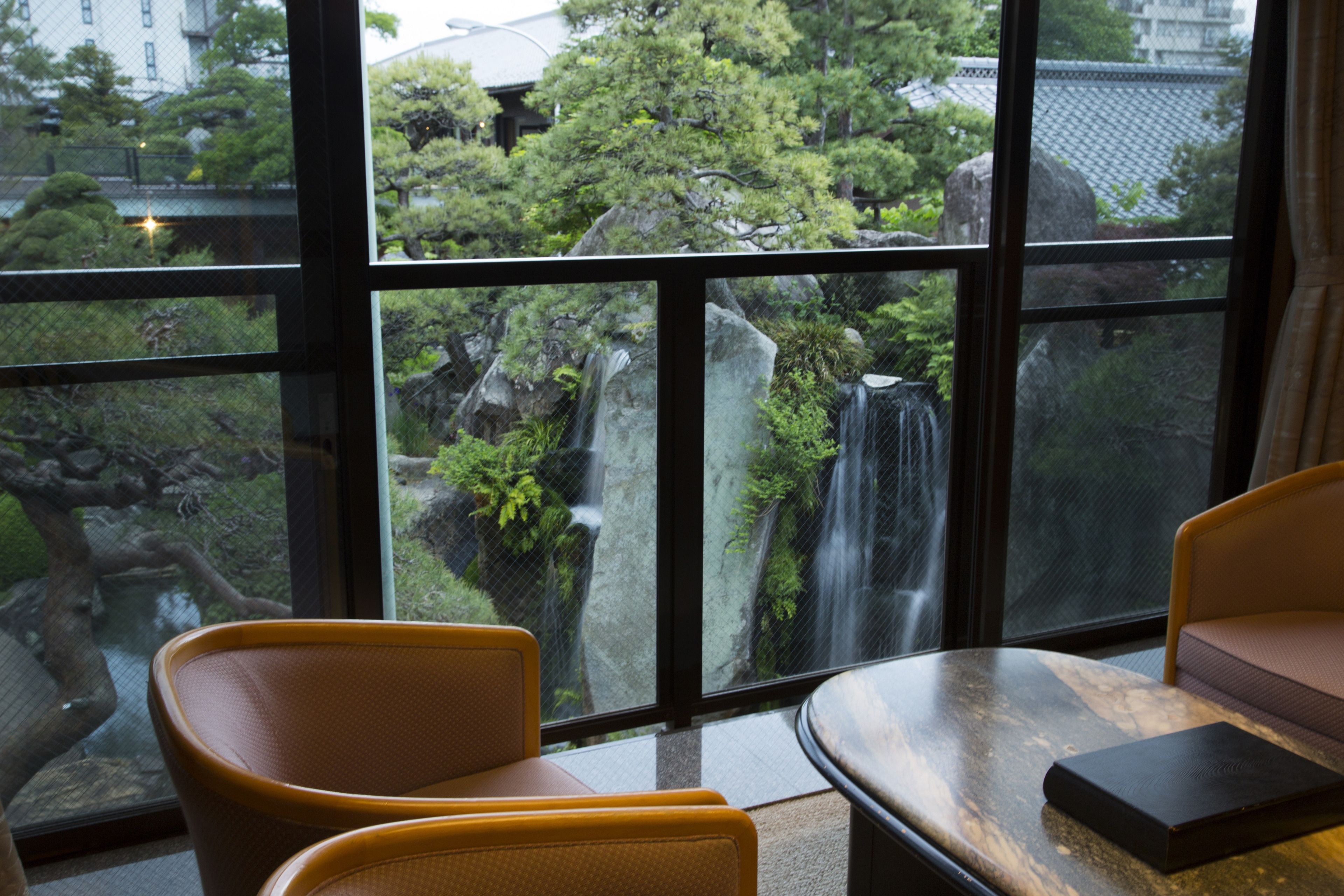 A room with a large window overlooking a waterfall and lush greenery. There are two chairs and a table in the room.