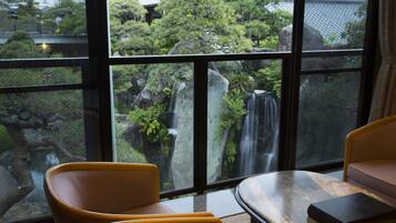 A room with a large window overlooking a waterfall and lush greenery. There are two chairs and a table in the room.