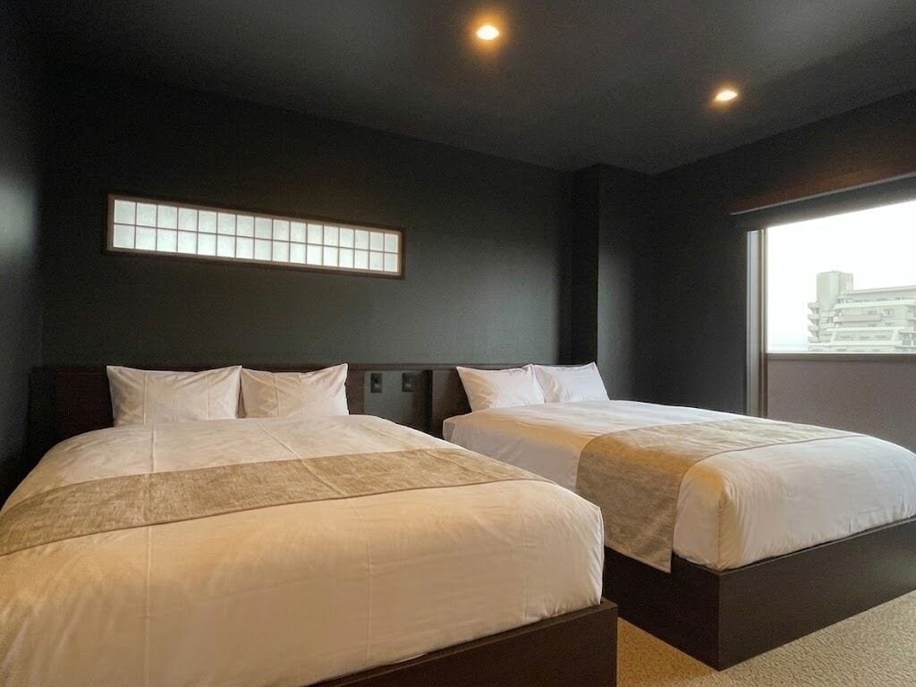 A hotel room with two beds, a large window, and a wall-mounted light fixture.